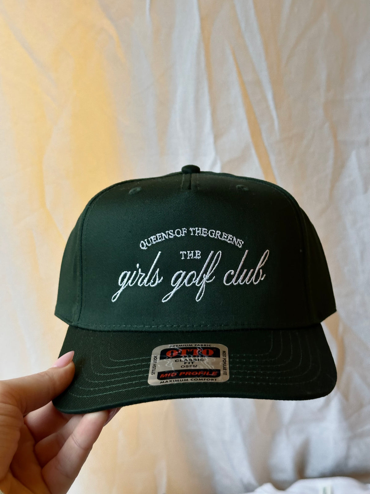 Golf trucker hat with embroidered “Girls Can Golf Too” and “golf club” lettering, displayed on turf with golf balls.