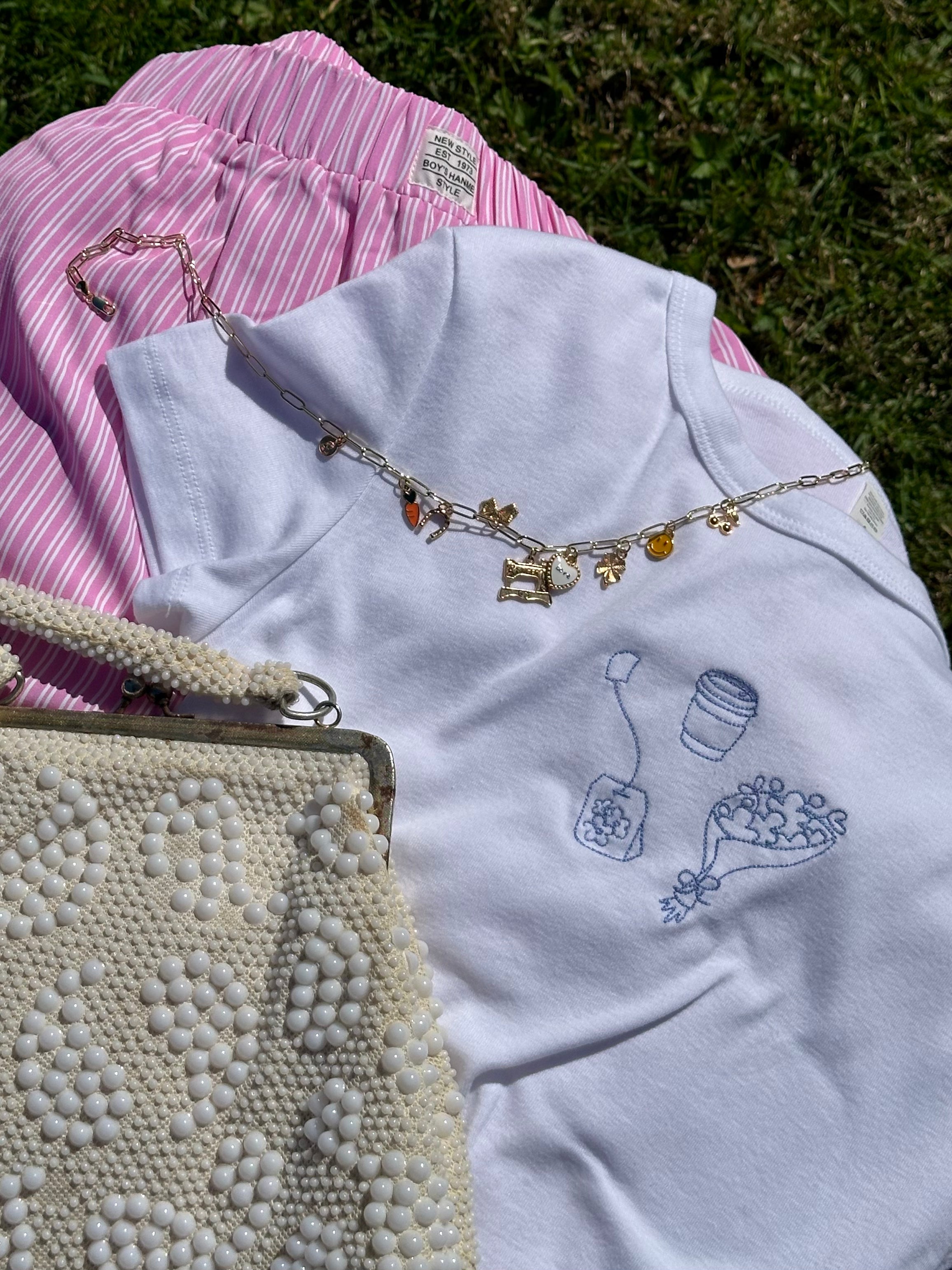 Flat lay of the Simple Saturdays embroidered baby tee styled with pink striped shorts, a charm necklace, and a beaded handbag.