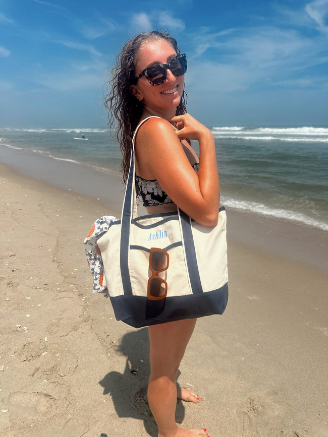 Model carrying a personalized canvas beach tote with navy trim and a custom embroidered name on the beach.