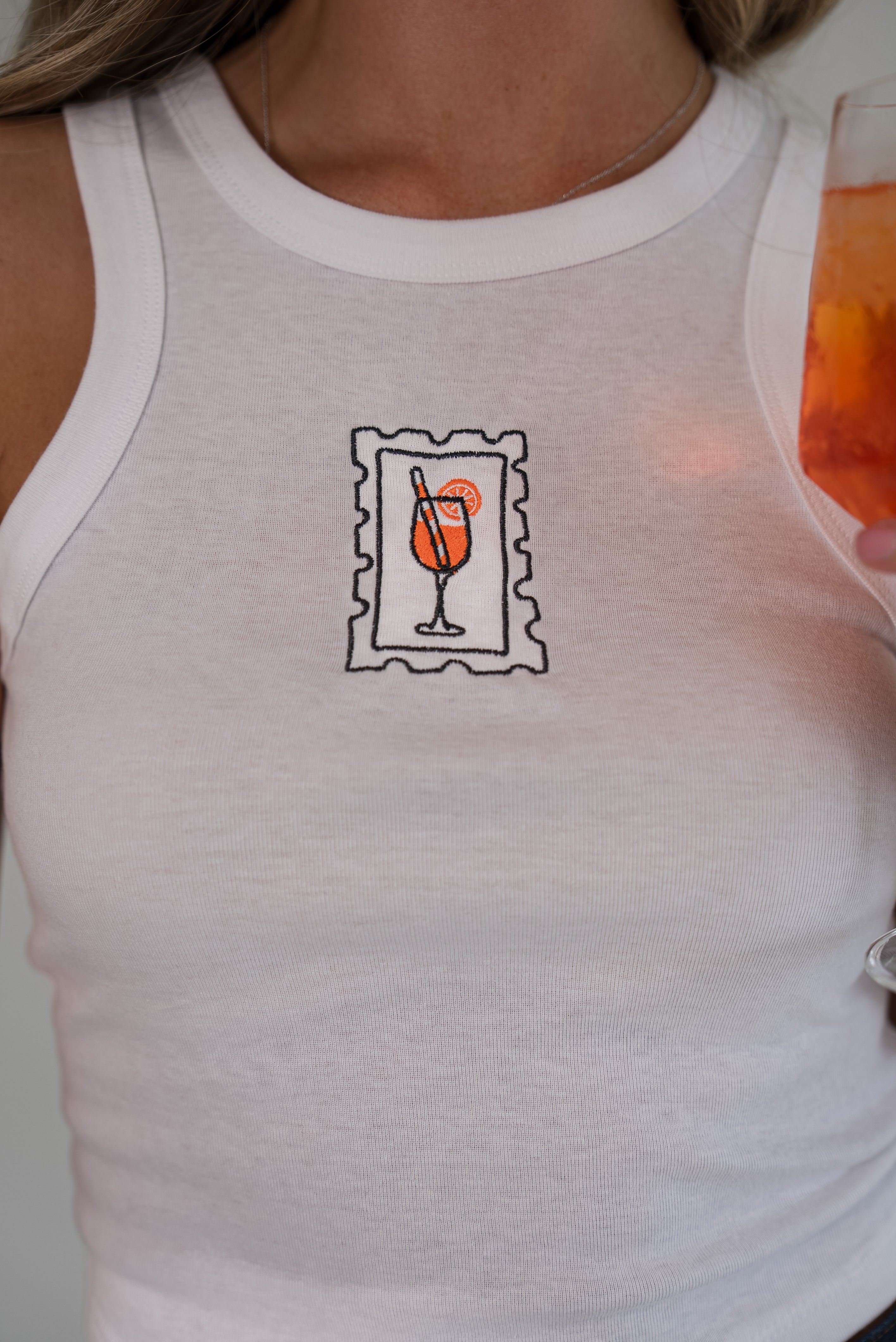 A close-up of a person wearing a white ribbed tank top with a small chest graphic of a cocktail glass inside a postage stamp outline, holding an orange drink.