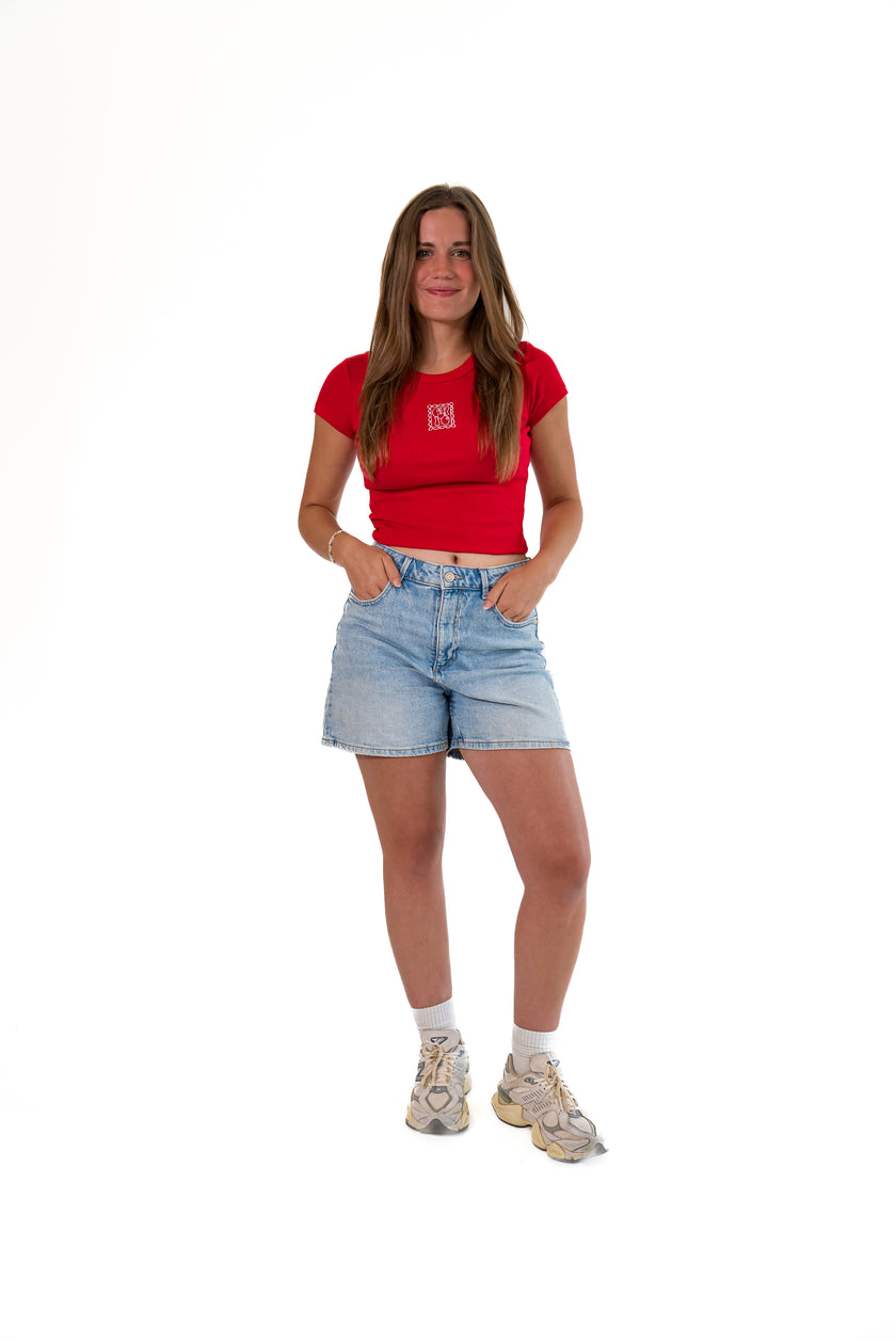 A woman modeling a red short-sleeve crop top with a small white square logo on the chest, paired with light-wash denim shorts, white socks, and beige sneakers, against a plain white background.