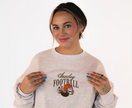 Model wearing an heather gray Sunday Football crewneck featuring embroidered football-inspired graphics including a helmet, football, and pom poms.