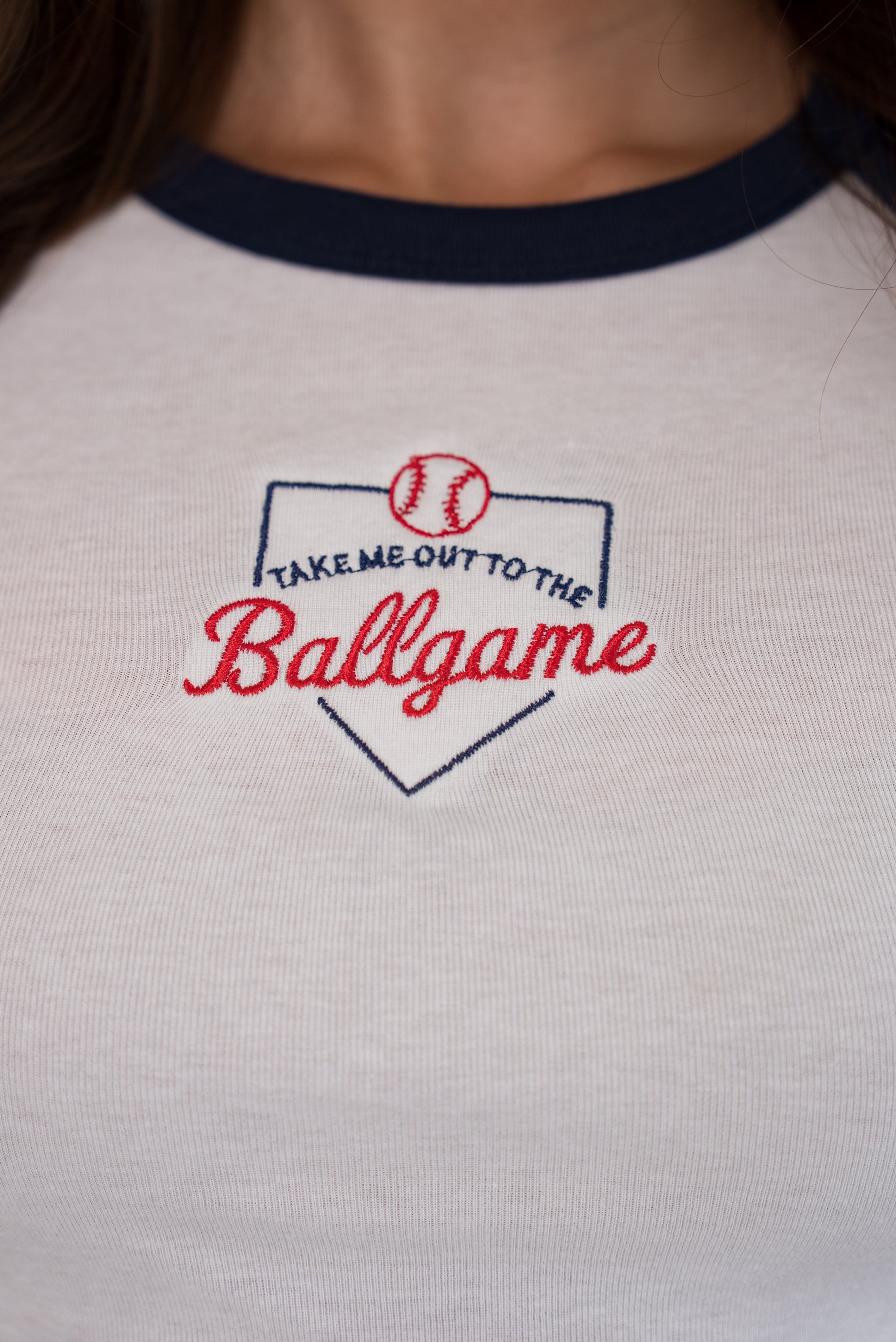 Close-up of the front of a white ringer tee with a navy collar, featuring a detailed embroidered home plate graphic with 