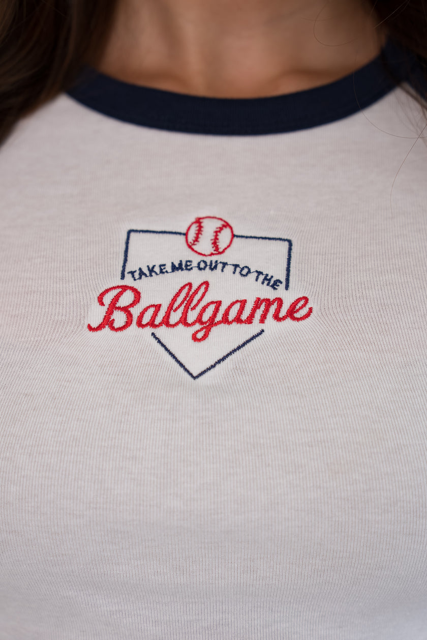 Close-up of the front of a white ringer tee with a navy collar, featuring a detailed embroidered home plate graphic with 