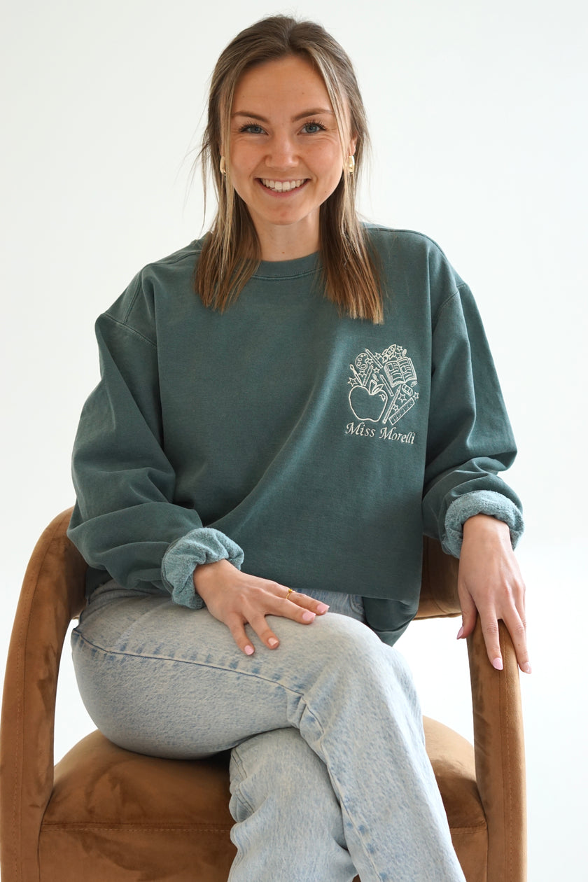 Model wearing a forest green personalized teacher crewneck sweatshirt with custom embroidered school-themed design and light wash jeans