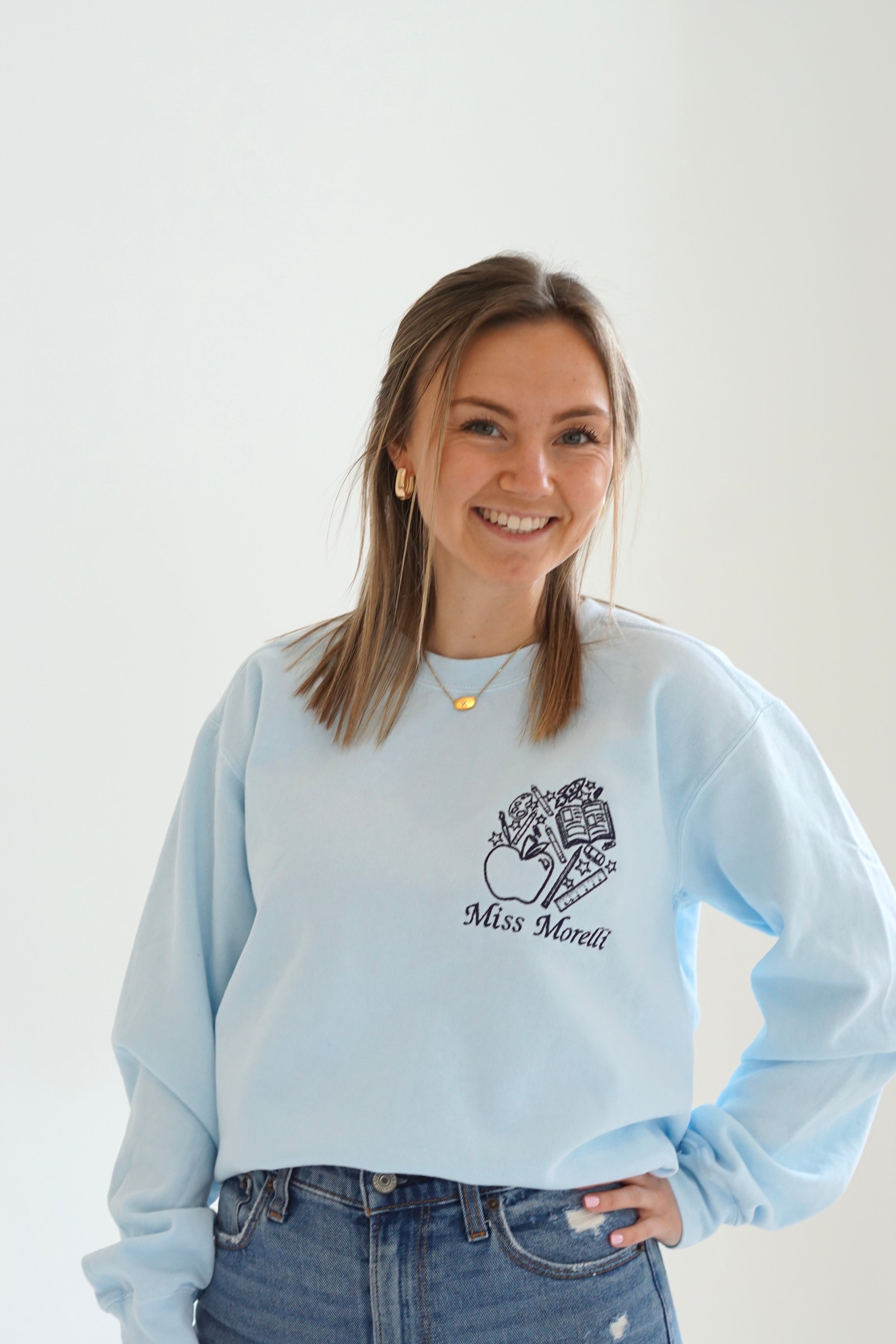 Close up of a light blue personalized teacher sweatshirt with custom embroidered name and school-themed artwork
