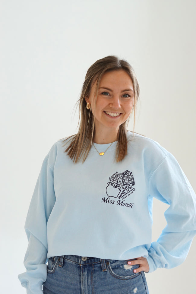 Close up of a light blue personalized teacher sweatshirt with custom embroidered name and school-themed artwork