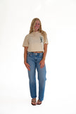 A woman modeling a beige short-sleeve crop top with a small black graphic on the left chest, paired with blue jeans and brown sandals, standing against a solid white background.