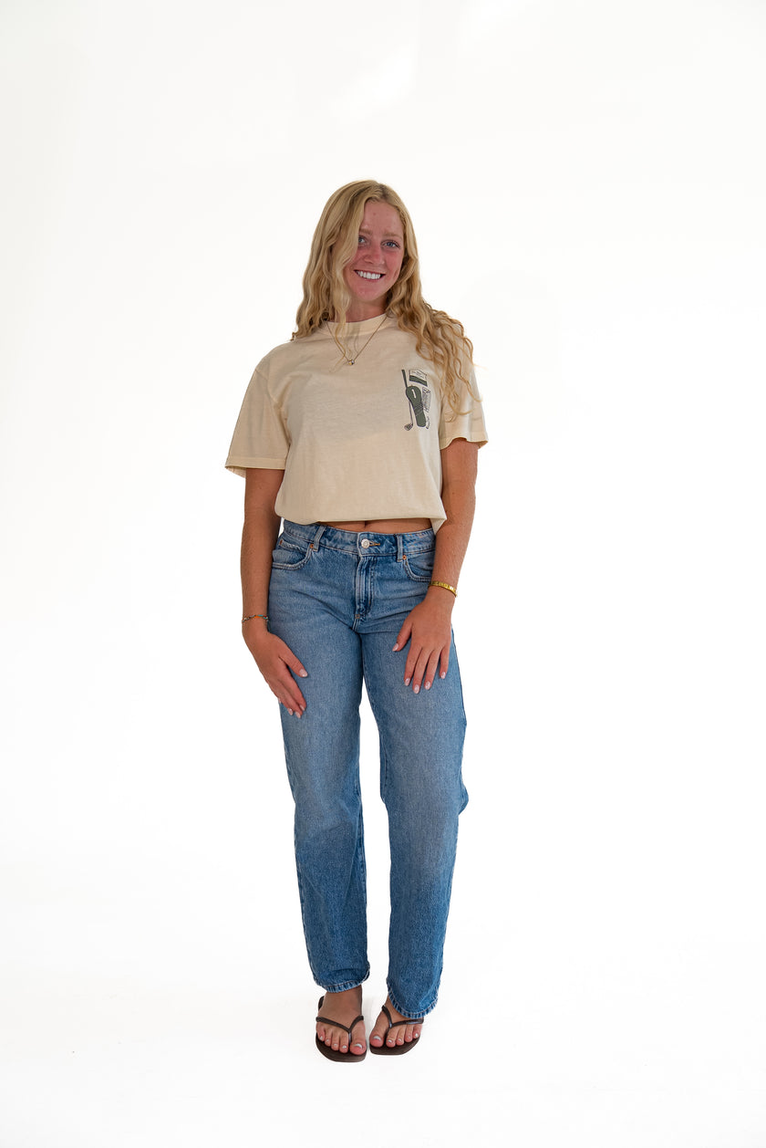A woman modeling a beige short-sleeve crop top with a small black graphic on the left chest, paired with blue jeans and brown sandals, standing against a solid white background.