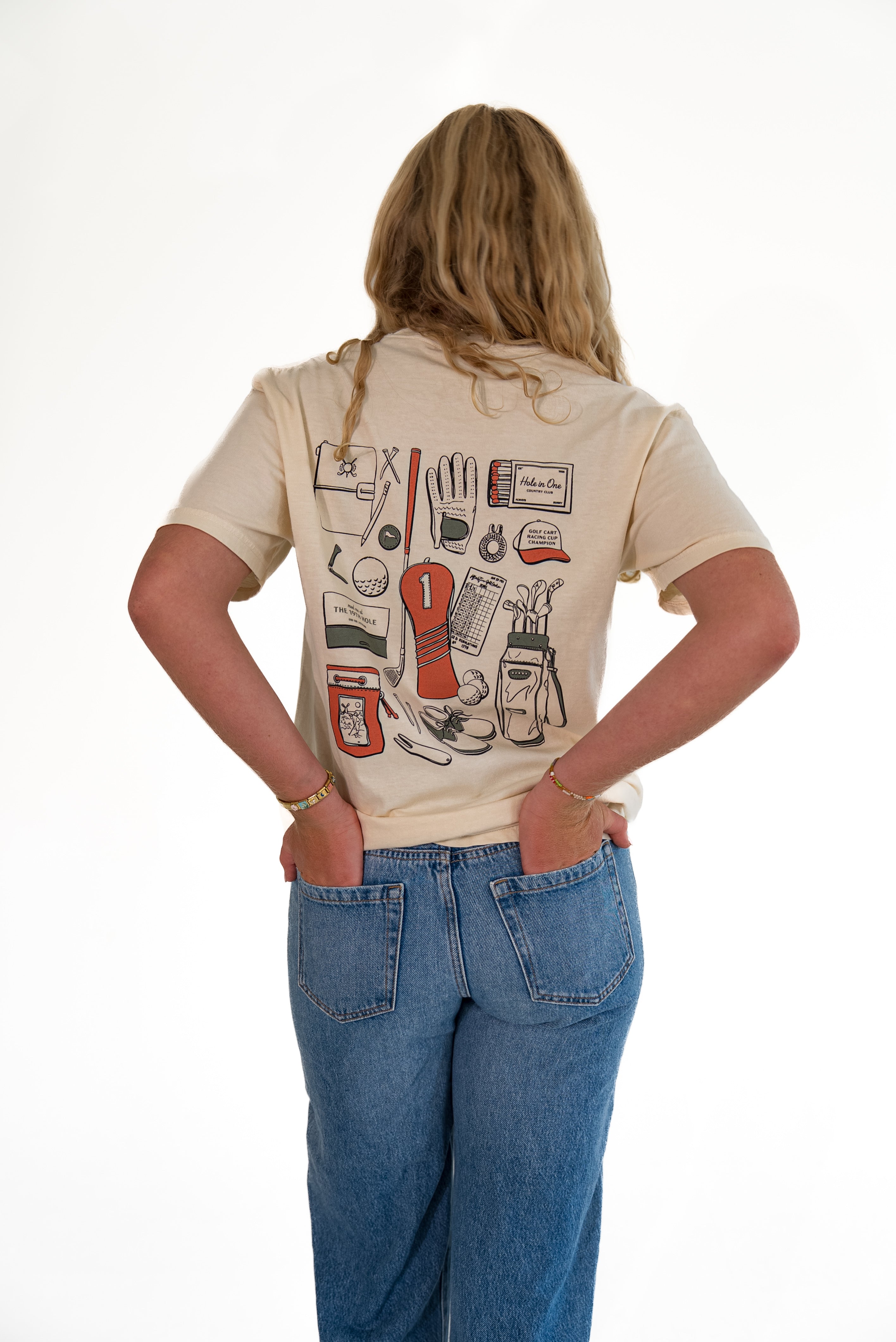 A woman viewed from behind wearing a cream-colored t-shirt with a vintage-style golf club graphic on the back, paired with blue jeans, against a plain white background.