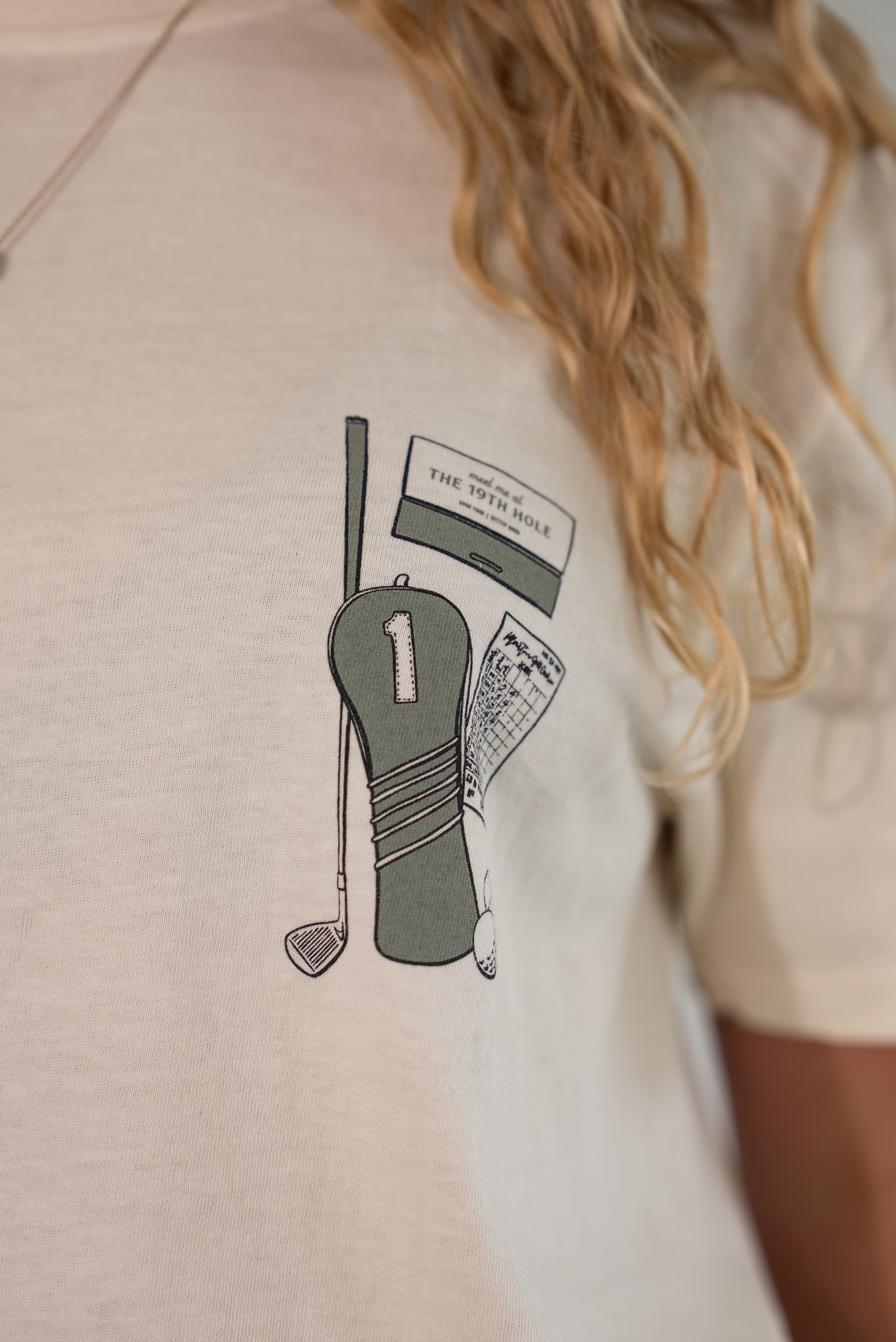 Close-up of a person wearing a light beige short-sleeve t-shirt with a graphic print of golf clubs and a ticket on the chest.
