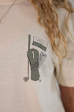 Close-up of a person wearing a light beige short-sleeve t-shirt with a graphic print of golf clubs and a ticket on the chest.