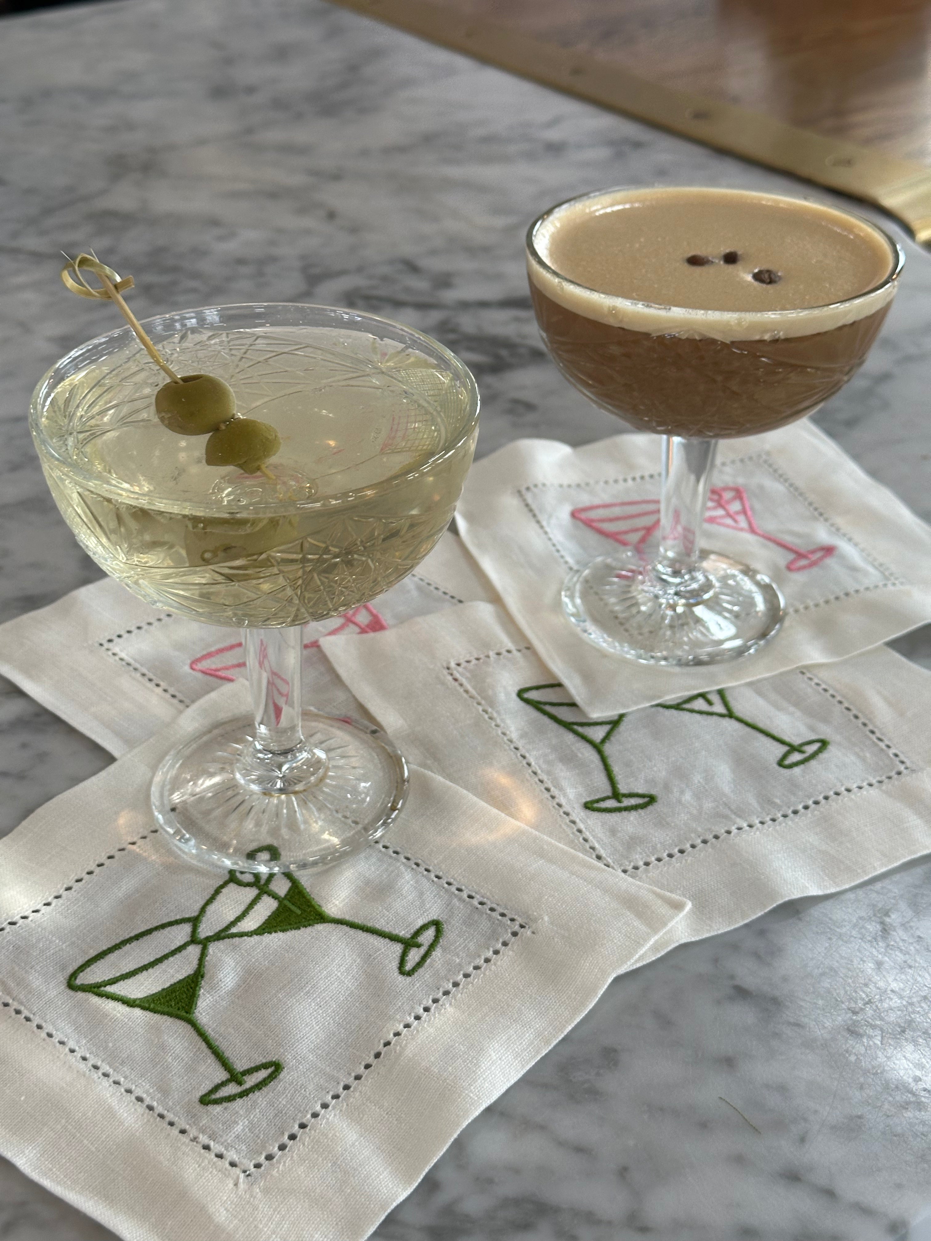 Embroidered martini cocktail napkins used as coasters beneath cocktails on a marble bar.