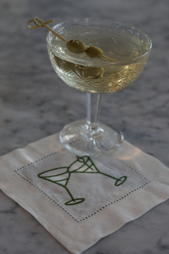 Embroidered martini cocktail napkin used as coasters beneath cocktail on a marble bar.