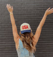 Cream and navy USA trucker hat with bold red embroidered lettering and a small American flag detail on the side.