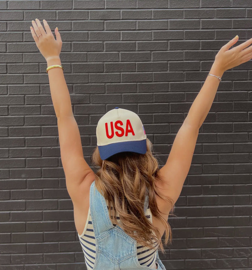 Cream and navy USA trucker hat with bold red embroidered lettering and a small American flag detail on the side.