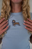 A close-up of a woman wearing a light blue t-shirt with an embroidered brown dachshund holding a blue bowtie on the chest.