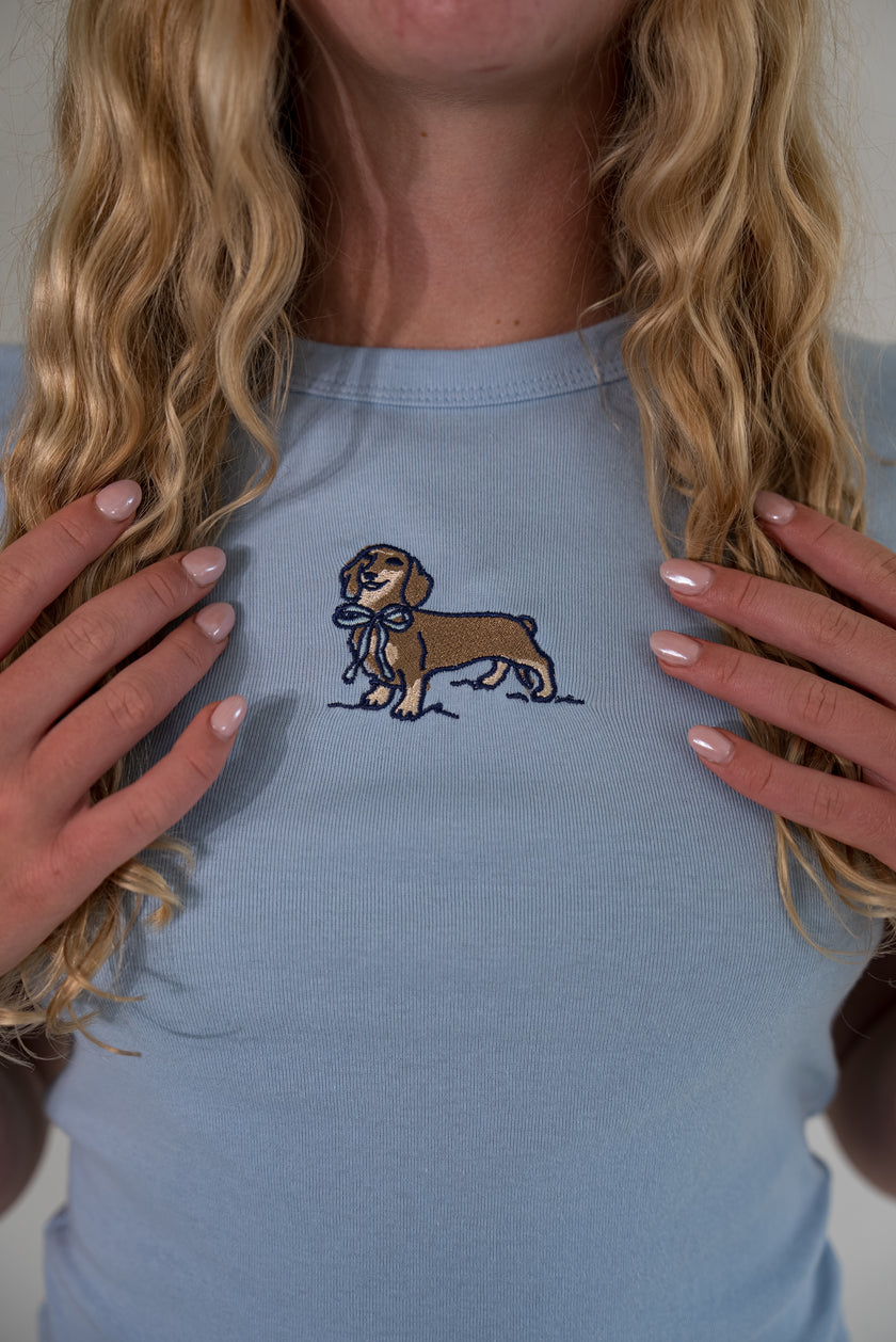 A close-up of a woman wearing a light blue t-shirt with an embroidered brown dachshund holding a blue bowtie on the chest.