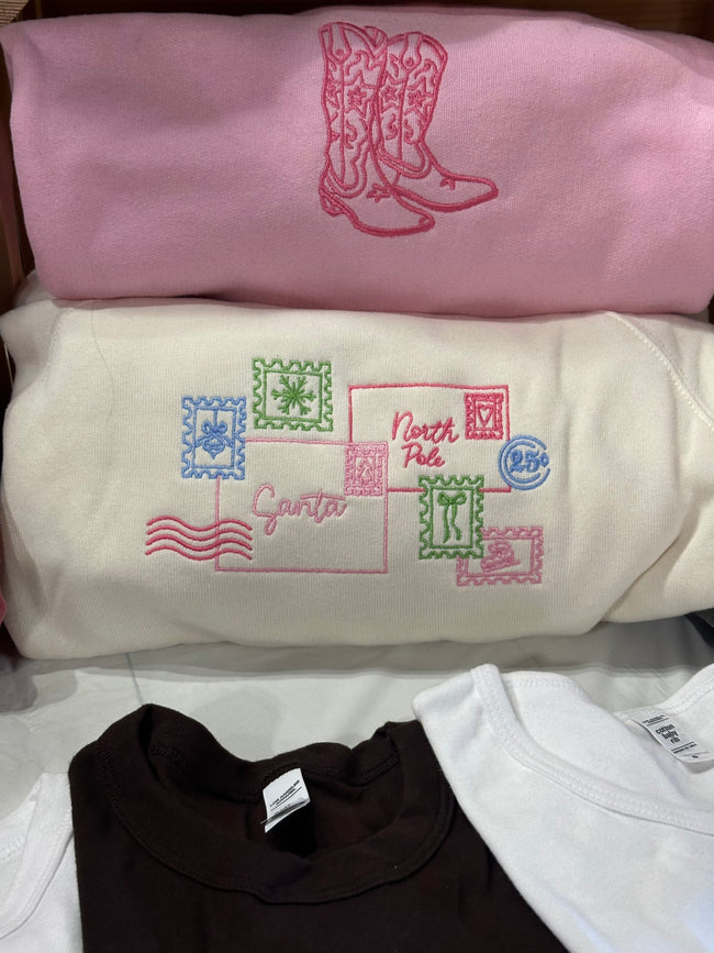 Folded Letter to Santa crewneck sweatshirts showcasing the embroidered holiday postcard design in multiple color options.
