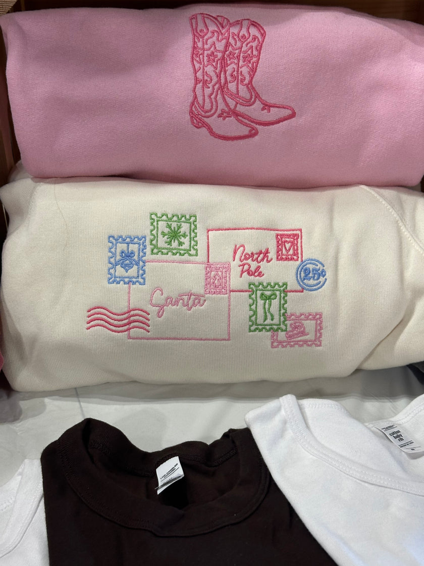 Folded Letter to Santa crewneck sweatshirts showcasing the embroidered holiday postcard design in multiple color options.