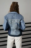Back view of a woman wearing a light wash color-block denim jacket with oversized fit and contrasting denim panels.